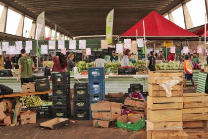 Pessoas fazem compras entre bancas de frutas e verduras em um mercado coberto de São Paulo.