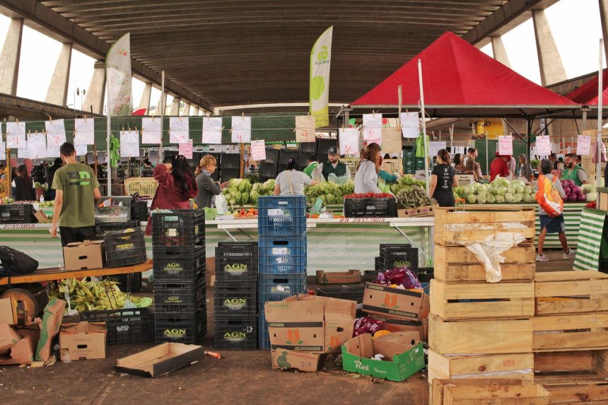 Pessoas fazem compras entre bancas de frutas e verduras em um mercado coberto de São Paulo.