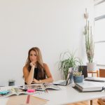 Empreendedora sorrindo à mesa de trabalho, em um escritório com plantas e materiais organizados.