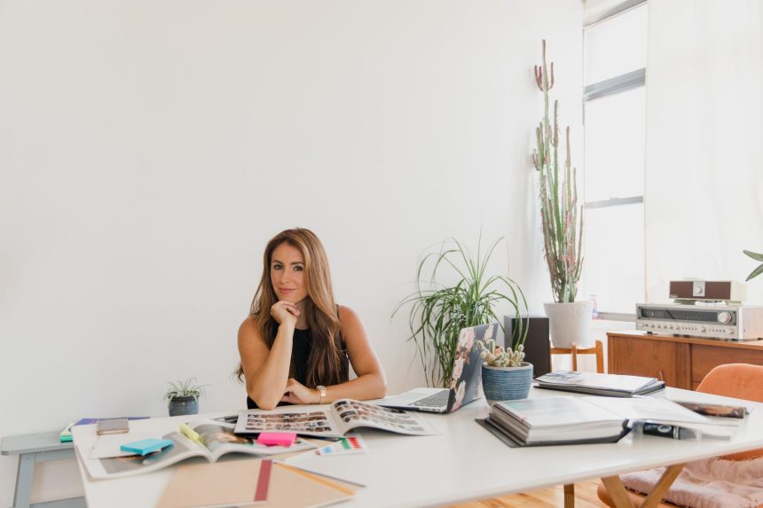 Empreendedora sorrindo à mesa de trabalho, em um escritório com plantas e materiais organizados.
