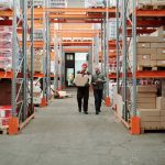Dois trabalhadores manuseiam uma encomenda em um amplo armazém cercado por prateleiras com caixas e produtos.