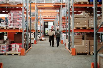 Dois trabalhadores manuseiam uma encomenda em um amplo armazém cercado por prateleiras com caixas e produtos.
