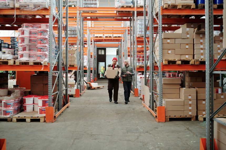 Dois trabalhadores manuseiam uma encomenda em um amplo armazém cercado por prateleiras com caixas e produtos.