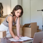Empreendedora administra seu negócio de roupas em um escritório doméstico, diante de um notebook e caixas de produtos.