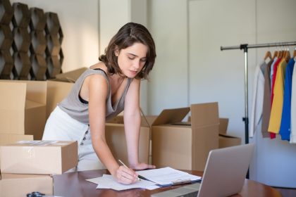 Empreendedora administra seu negócio de roupas em um escritório doméstico, diante de um notebook e caixas de produtos.