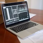 Notebook com gráficos na tela ao lado de caixas de papelão sobre uma mesa de madeira.