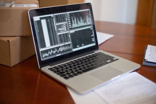 Notebook com gráficos na tela ao lado de caixas de papelão sobre uma mesa de madeira.