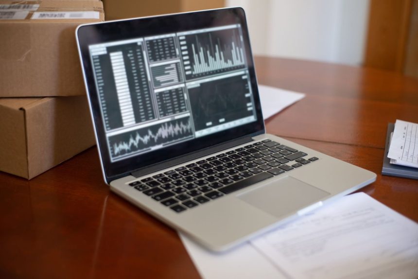 Notebook com gráficos na tela ao lado de caixas de papelão sobre uma mesa de madeira.