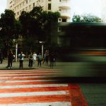Pessoas aguardam para atravessar uma rua enquanto um ônibus passa desfocado em uma via urbana movimentada.