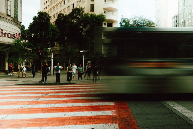 Pessoas aguardam para atravessar uma rua enquanto um ônibus passa desfocado em uma via urbana movimentada.