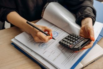 Profissional de contabilidade usa uma calculadora e assina documentos financeiros em uma mesa de trabalho.