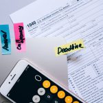 Mesa de escritório com documentos fiscais, celular exibindo uma calculadora e anotações ao lado de clipes de papel