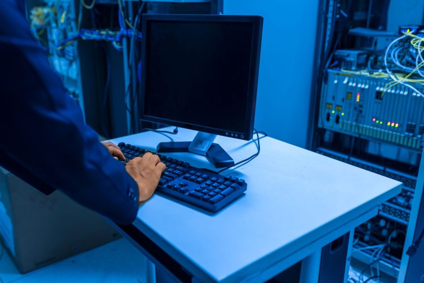 Profissional de tecnologia opera um computador em uma sala de servidores, gerenciando sistemas de rede e dispositivos conectados.
