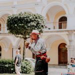 Artista de rua toca maracas em uma praça ensolarada diante de um edifício histórico.