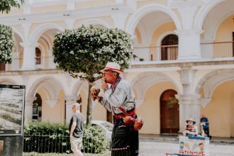 Artista de rua toca maracas em uma praça ensolarada diante de um edifício histórico.