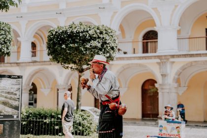 Artista de rua toca maracas em uma praça ensolarada diante de um edifício histórico.