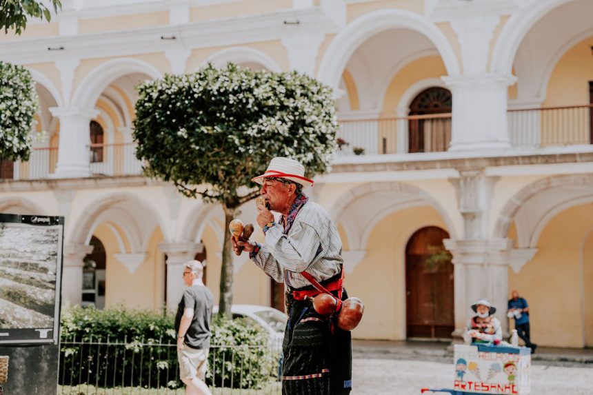 Artista de rua toca maracas em uma praça ensolarada diante de um edifício histórico.