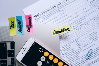 Mesa de escritório com documentos fiscais, celular exibindo uma calculadora e anotações ao lado de clipes de papel