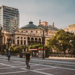 Turistas e pedestres caminham por uma praça no centro de São Paulo, diante de edifícios modernos e do Theatro Municipal.