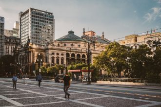 Turistas e pedestres caminham por uma praça no centro de São Paulo, diante de edifícios modernos e do Theatro Municipal.