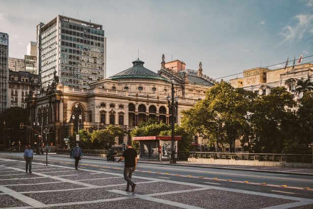 Turistas e pedestres caminham por uma praça no centro de São Paulo, diante de edifícios modernos e do Theatro Municipal.